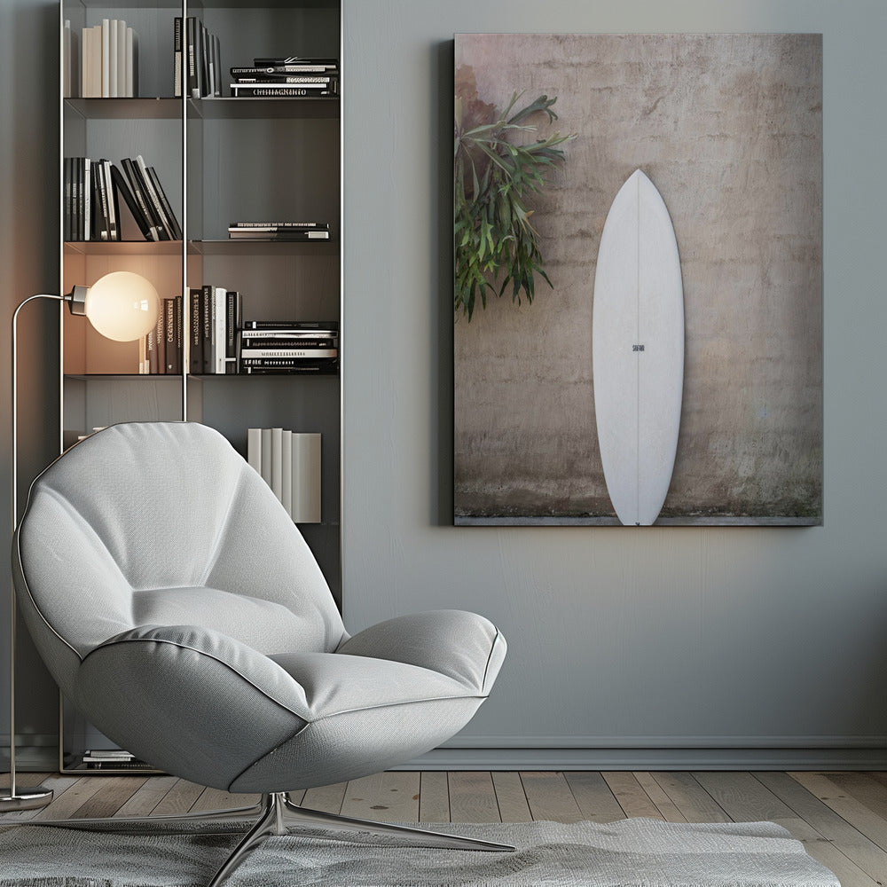 A minimalist photograph of a white surfboard leaning against a textured beige concrete wall. To the left, a large green staghorn fern adds a touch of nature. The entire scene is enclosed in a simple black frame. Artwork
