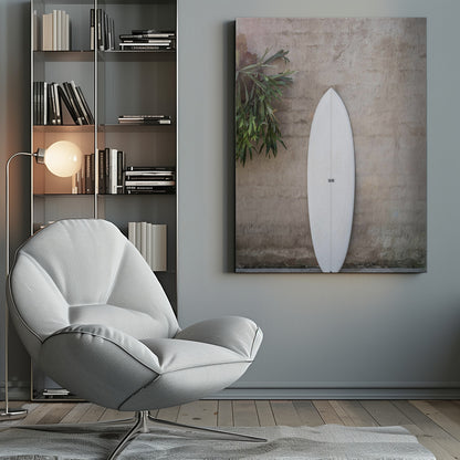 A minimalist photograph of a white surfboard leaning against a textured beige concrete wall. To the left, a large green staghorn fern adds a touch of nature. The entire scene is enclosed in a simple black frame. Artwork