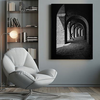 A dramatic black and white photograph of a person walking through a long brick arcade. Sunlight creates strong, contrasting shadows, highlighting the repeating arches that recede into the darkness, creating a powerful sense of depth and perspective. Decor