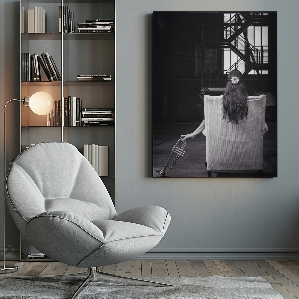A black and white photograph of a woman seen from behind, sitting in a large, light-colored corduroy armchair. She has long, dark hair with a white flower pinned in it. Her left hand holds a trumpet, resting it on the dark floor of what appears to be an industrial warehouse with a metal staircase in the background. Wall Art