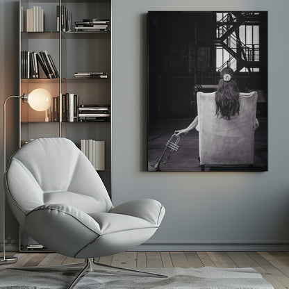 A black and white photograph of a woman seen from behind, sitting in a large, light-colored corduroy armchair. She has long, dark hair with a white flower pinned in it. Her left hand holds a trumpet, resting it on the dark floor of what appears to be an industrial warehouse with a metal staircase in the background. Wall Art
