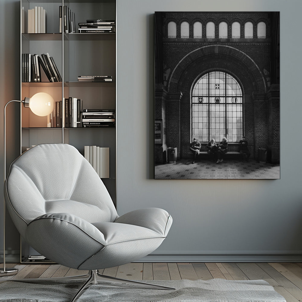 A dramatic black and white photograph of a large, brick-walled waiting area, possibly a train station. Four people are seated on a long bench beneath a massive arched window, which dominates the scene. The lighting creates a moody, contemplative atmosphere, highlighting the grandeur of the architecture. Artwork