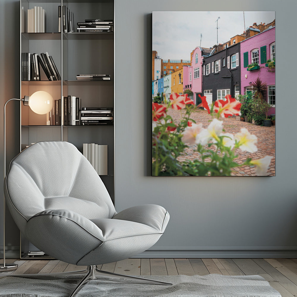 A low-angle view of a charming, narrow cobblestone street lined with colorful row houses in shades of pink, black, yellow, and blue, with blurred red and white petunias in the foreground. Wall Art