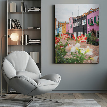 A low-angle view of a charming, narrow cobblestone street lined with colorful row houses in shades of pink, black, yellow, and blue, with blurred red and white petunias in the foreground. Wall Art