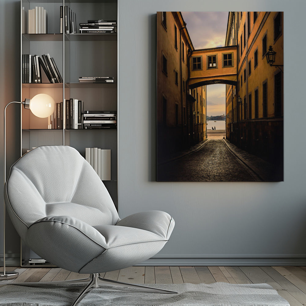 A perspective view down a narrow, shadowy cobblestone alley in an old European city, framed by warm, ochre-colored buildings. An arched walkway connects the buildings overhead, and the alley opens to a view of a tranquil body of water at sunset, with two small figures silhouetted by the shore. Decor