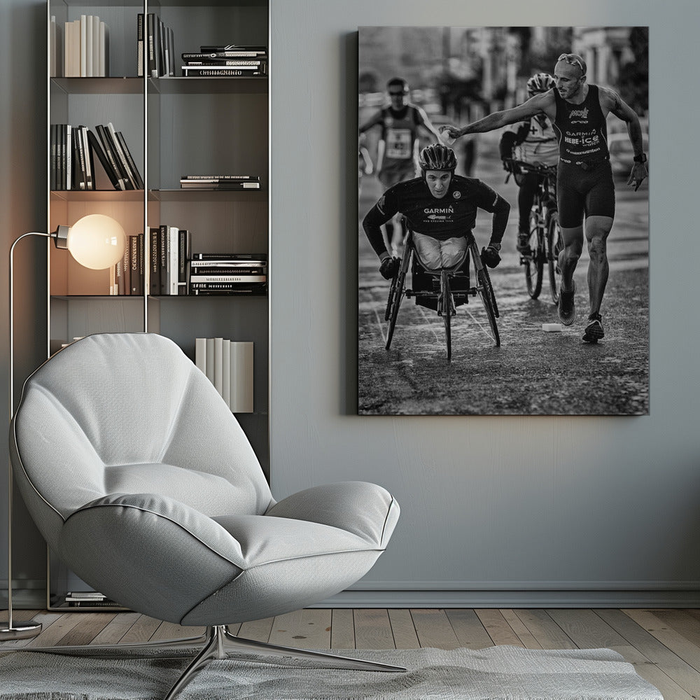 A dynamic black and white action photograph of a para-athlete in a racing wheelchair during a race. A running athlete beside him reaches out to hand him a sponge, showcasing a moment of camaraderie and support. Other competitors are blurred in the background on the wet asphalt. Print