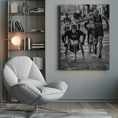 A dynamic black and white action photograph of a para-athlete in a racing wheelchair during a race. A running athlete beside him reaches out to hand him a sponge, showcasing a moment of camaraderie and support. Other competitors are blurred in the background on the wet asphalt. Print
