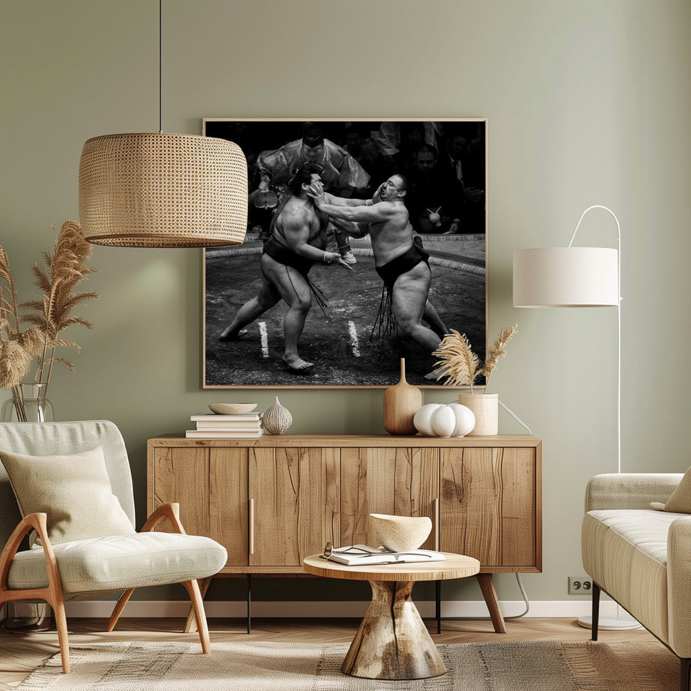 A dynamic black and white action photograph of a sumo wrestling match. Two large wrestlers are in the middle of a clay ring, one pushing forcefully against the other's face. A referee in traditional robes stands behind them, and spectators watch from the background. The entire scene is captured within a light wood frame. Artwork