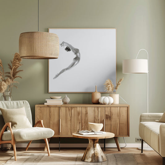 A minimalist black and white photograph of a graceful dancer frozen in mid-air, her body arched backward in an elegant C-shape against a stark white background. The artwork is presented in a light wood frame. Decor