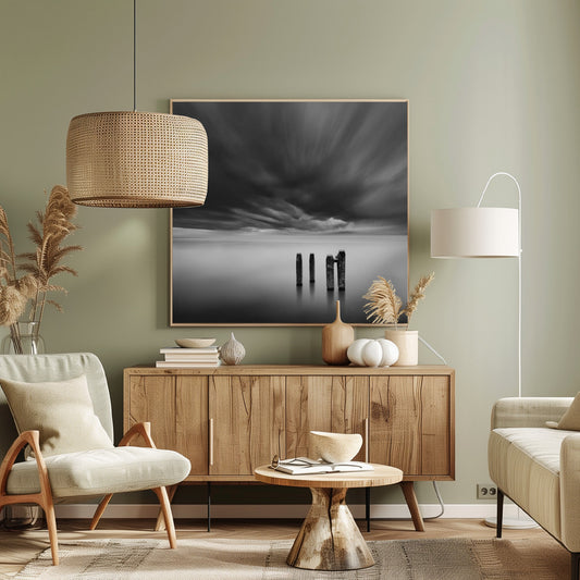 A framed, square, black and white long-exposure photograph of a minimalist seascape. Four dark wooden pilings emerge from still, misty water under a dramatic, streaked sky of dark clouds. Wall Art