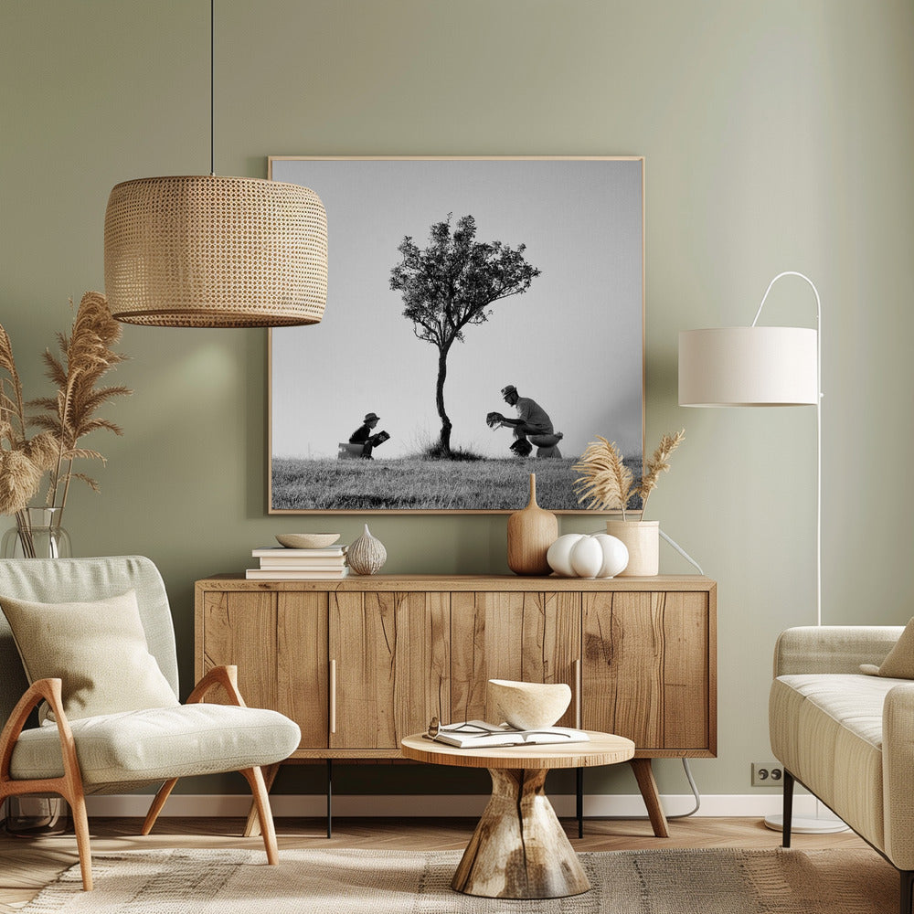 A framed, black and white photograph depicting a surreal scene of a man and a child sitting on toilets in an open grassy field under a single tree, both wearing hats and reading. Print