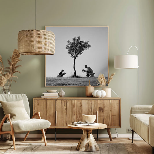 A framed, black and white photograph depicting a surreal scene of a man and a child sitting on toilets in an open grassy field under a single tree, both wearing hats and reading. Print