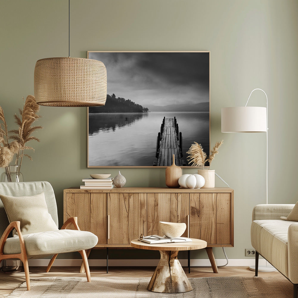 A framed, moody black and white photograph of a wooden pier extending into a calm lake under a cloudy sky, with a forested shoreline and distant mountains in the background. Poster