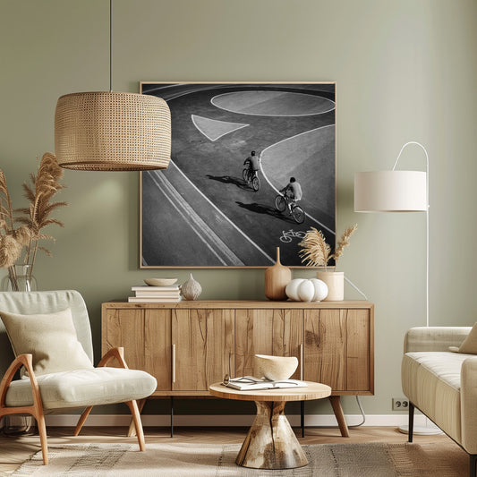 A framed, high-angle black and white photograph of two people cycling on a curving asphalt bike path. The path is marked with bold white lines, including a large arrow and a bicycle symbol, creating a strong graphic pattern. The cyclists cast long shadows on the pavement. Poster