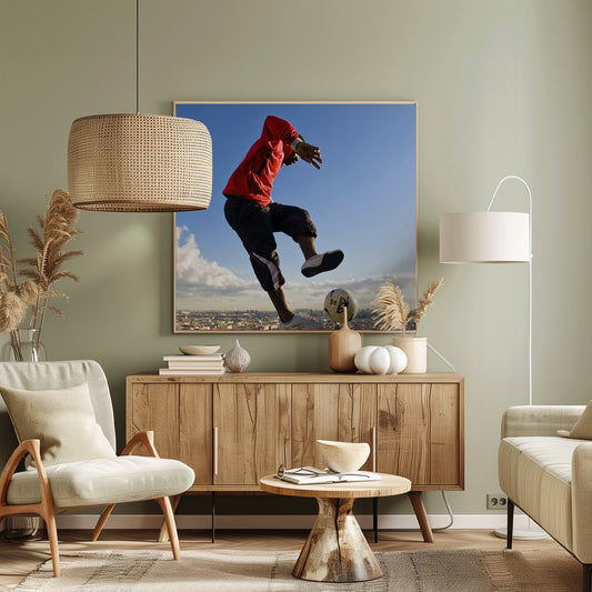 A dynamic low-angle shot of a person in a red jacket and black pants skillfully juggling a soccer ball, suspended in mid-air high above a sprawling city skyline under a clear blue sky, all enclosed within a light wood frame. Artwork