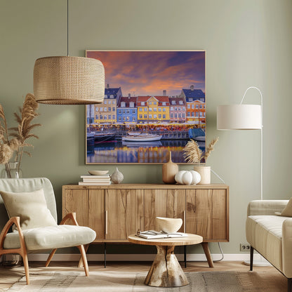 A framed photograph of the Nyhavn harbor in Copenhagen at dusk, featuring a row of brightly colored historic buildings along the canal. Sailboats are moored in the water, which reflects the warm glow of the building lights and the vibrant orange and purple sunset sky. Print