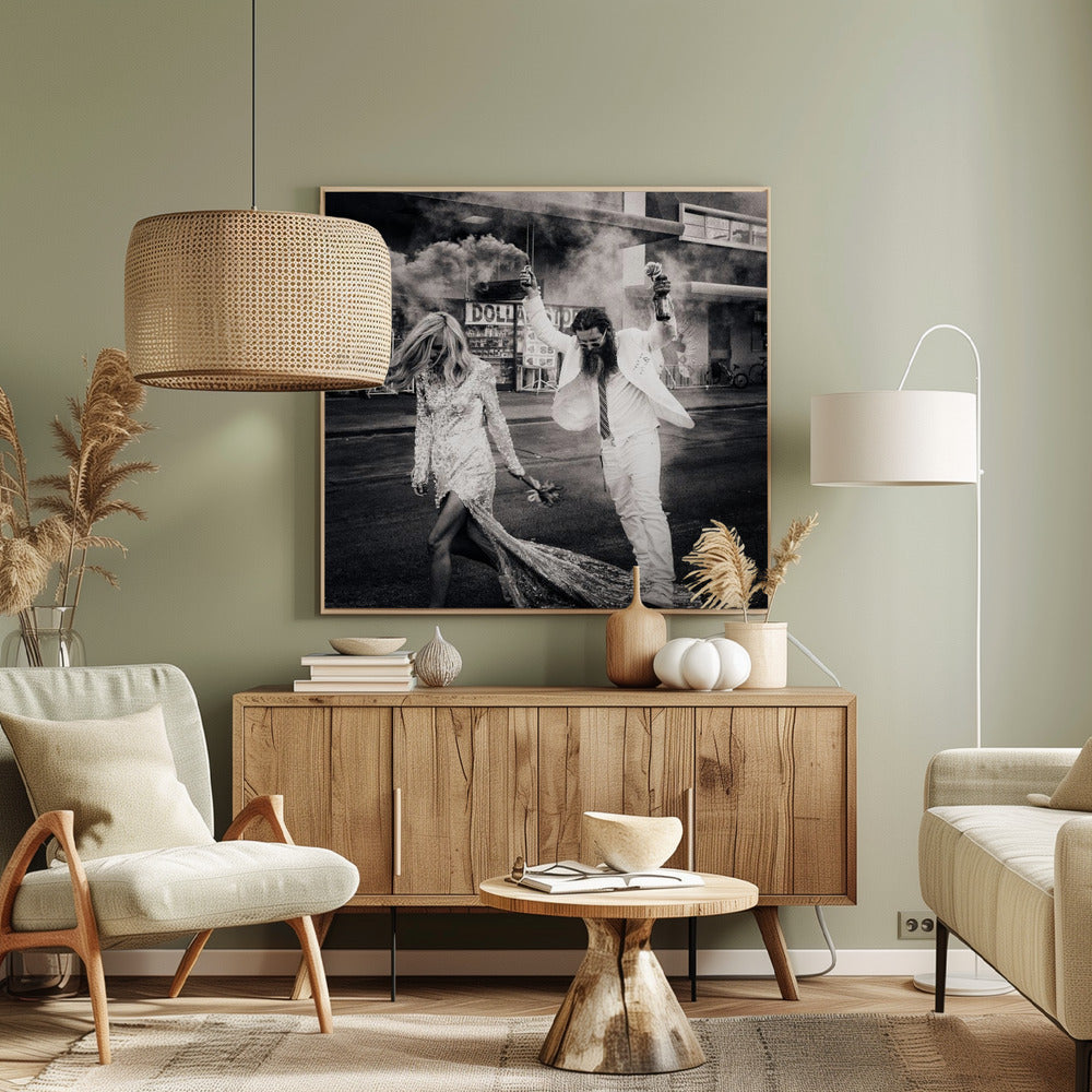 A black and white photograph in a light wood frame showing a stylish couple walking down a street. The woman wears a sparkling sequin gown, and the man, in a white suit with a long beard, triumphantly raises his arms, holding a smoke bomb and a bottle with a rose. Decor