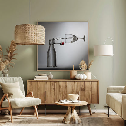 A conceptual photograph in a light wood frame depicting water pouring horizontally from an open swing-top glass bottle into a floating, sideways wine glass against a simple gray background, capturing the liquid in mid-air. Print