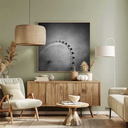 A moody, black and white long-exposure photograph of a large Ferris wheel, creating a soft, blurry effect of its rotation against a grainy gray sky. The artwork is presented in a simple, light wood frame. Poster