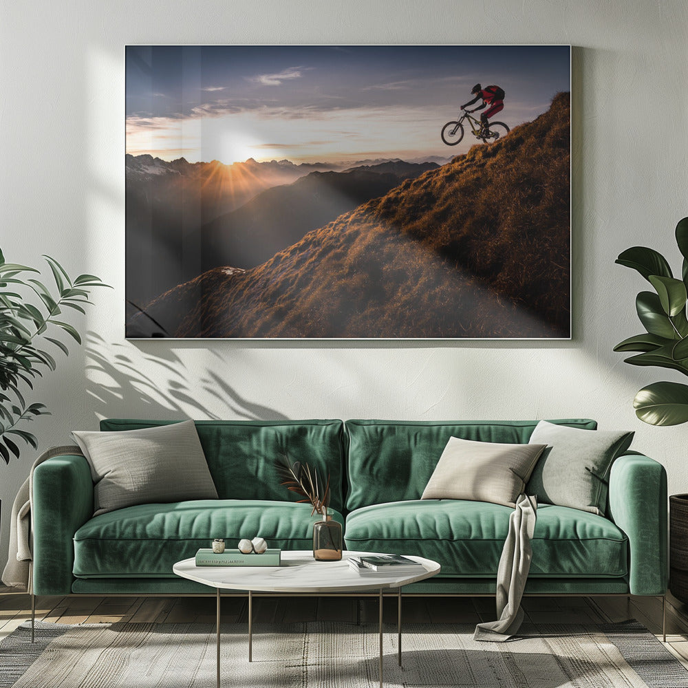 A mountain biker in mid-air, jumping down a steep grassy mountain ridge at sunrise. The sun is low on the horizon, casting a golden glow and long sunbeams over a vast, layered mountain range in the background. Poster