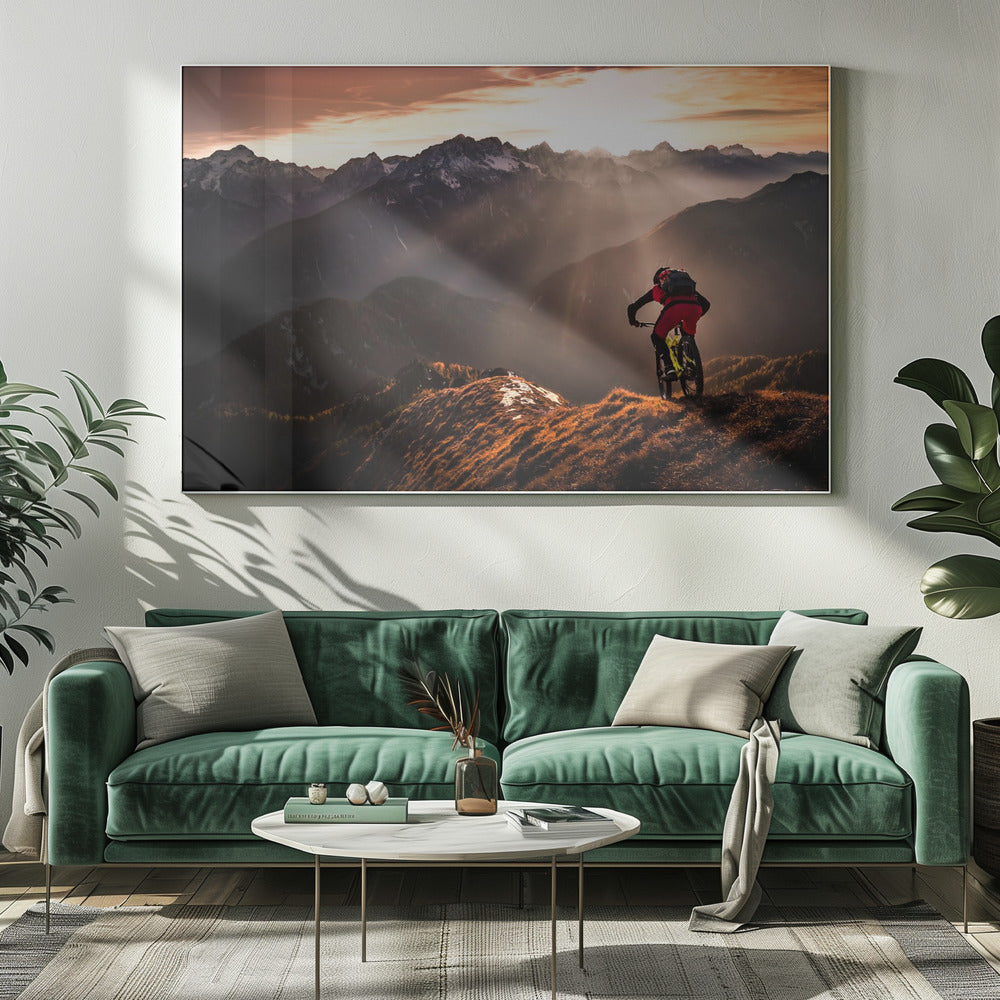A mountain biker in red and black gear pauses on a grassy, sunlit ridgeline, looking out at a vast mountain range with snow-capped peaks as the sun sets behind them, casting dramatic rays of light through the valleys. Poster