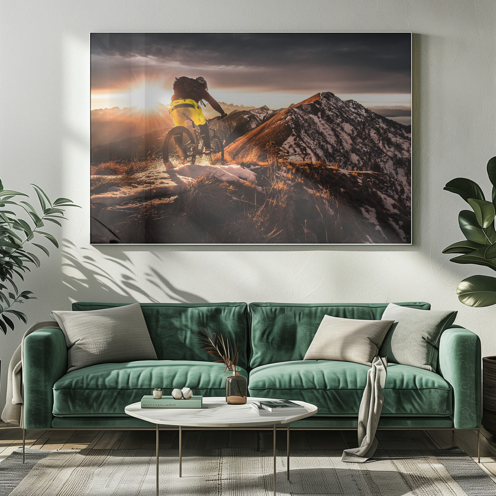 A mountain biker seen from behind rides along a narrow, snowy trail on a mountain ridge during a dramatic sunset, with the sun flaring brightly and a vast, snow-capped mountain range in the background. Decor