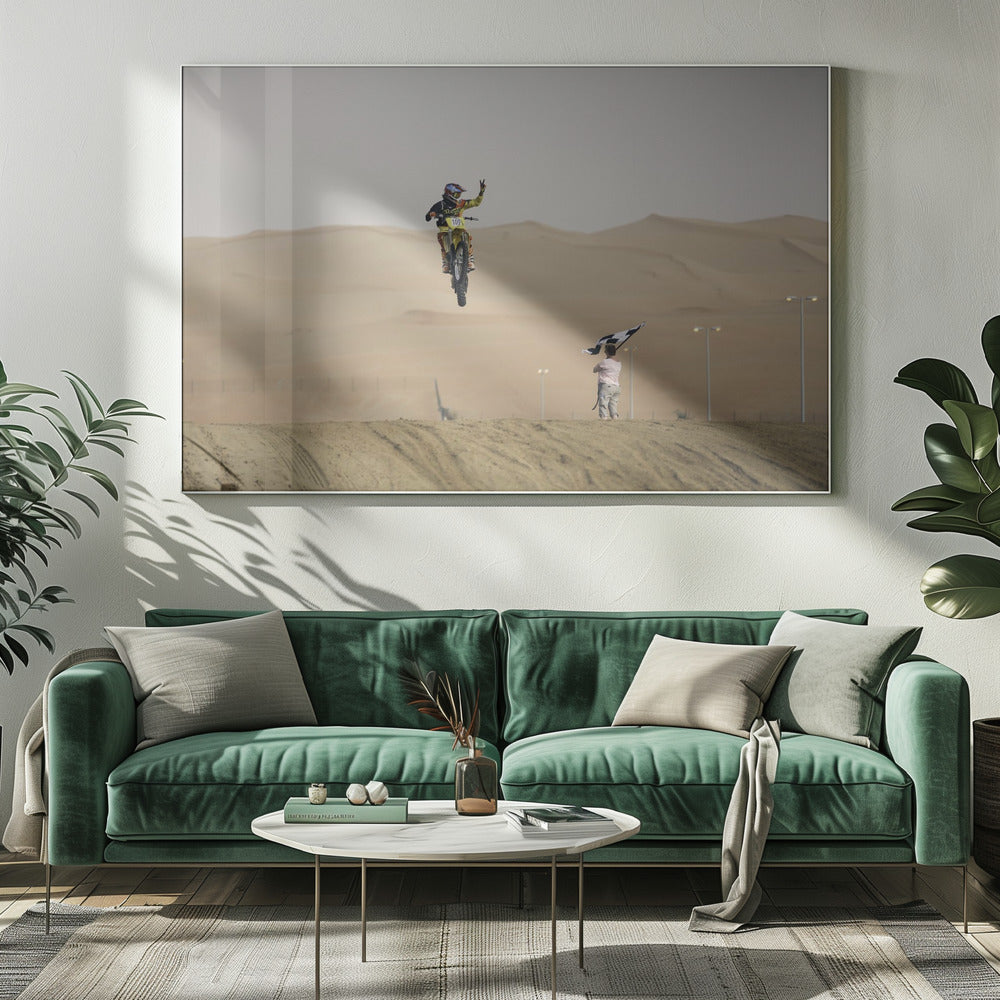 A motocross rider on a yellow dirt bike is captured mid-air during a jump in a desert landscape, flashing a peace sign with their left hand. In the background, rolling sand dunes stretch under a hazy sky, and to the right, a person on a hill waves a black and white checkered flag. Print