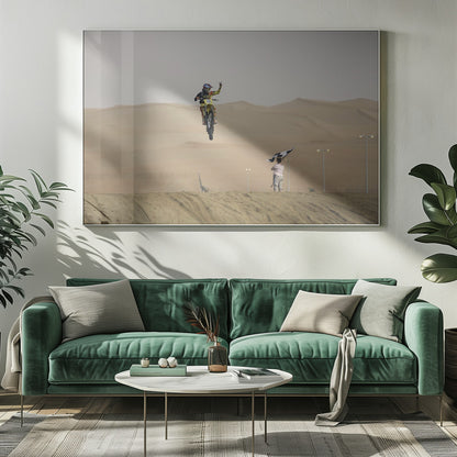 A motocross rider on a yellow dirt bike is captured mid-air during a jump in a desert landscape, flashing a peace sign with their left hand. In the background, rolling sand dunes stretch under a hazy sky, and to the right, a person on a hill waves a black and white checkered flag. Print