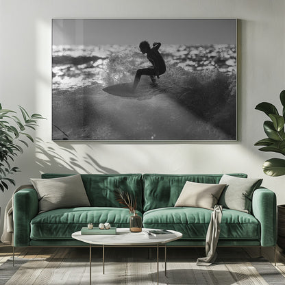 A black and white action shot of a surfer silhouetted against the sparkling ocean. The surfer is crouched low on their board, carving through a wave and creating a dramatic spray of water. The photo is enclosed in a silver frame. Print