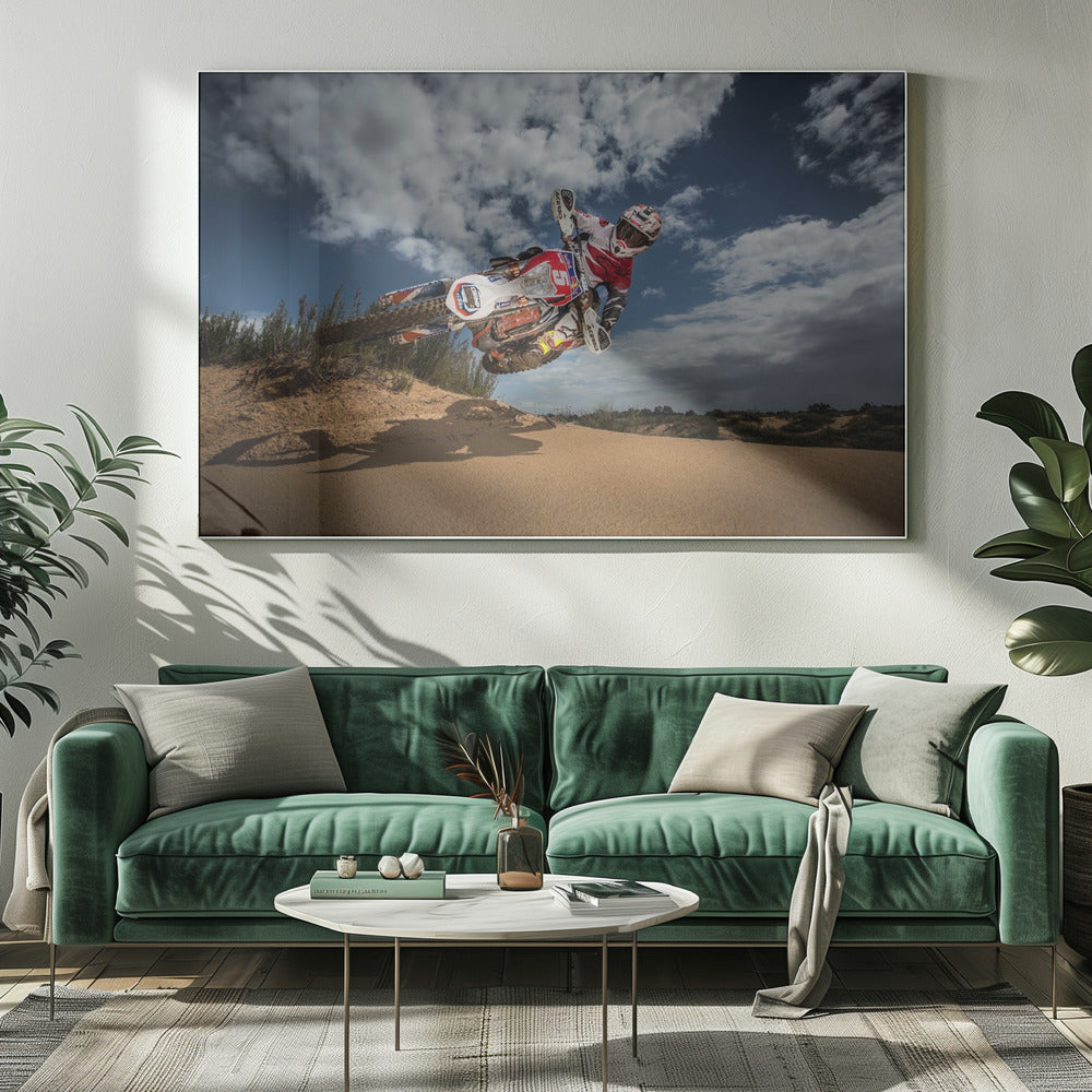 A low-angle action shot of a motocross rider on a dirt bike catching major air over a sand dune against a cloudy blue sky. The image is presented within a silver frame. Decor