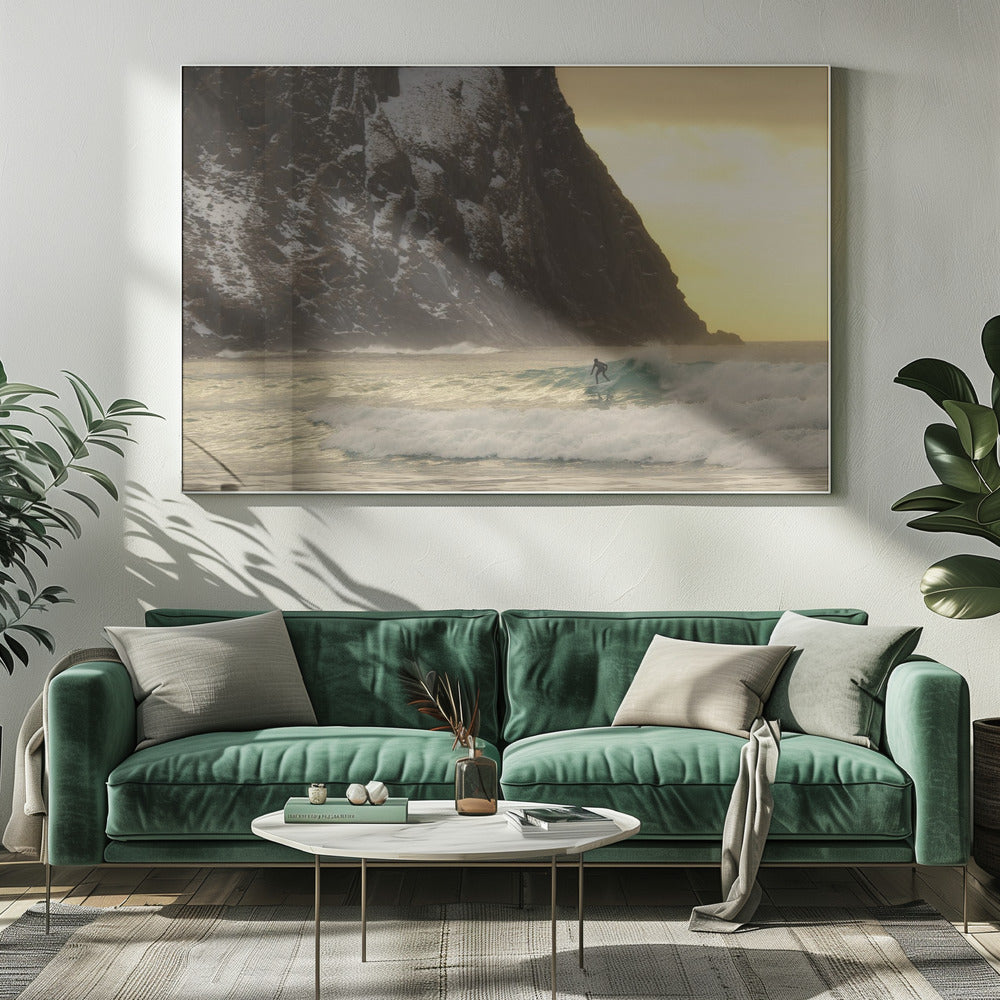 A framed photograph captures a lone surfer in a black wetsuit riding a turquoise wave. In the background, a massive, steep, snow-dusted mountain looms over the cold sea under a pale yellow, overcast sky. Print