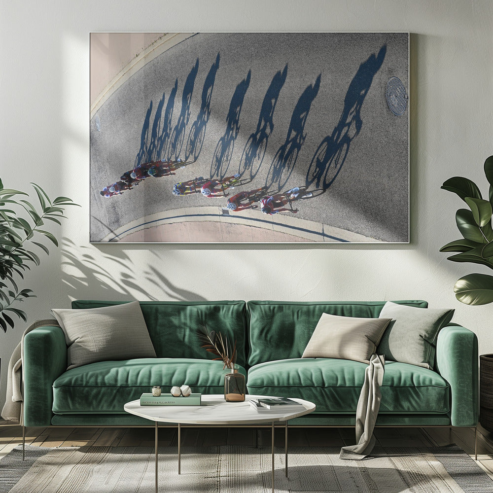 An aerial, high-angle photograph of a line of cyclists racing on a curved asphalt road. The bright sun casts long, dark, dramatic shadows of the riders and their bicycles, creating a striking diagonal pattern across the pavement. The image is framed in silver. Print