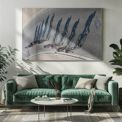 An aerial, high-angle photograph of a line of cyclists racing on a curved asphalt road. The bright sun casts long, dark, dramatic shadows of the riders and their bicycles, creating a striking diagonal pattern across the pavement. The image is framed in silver. Print