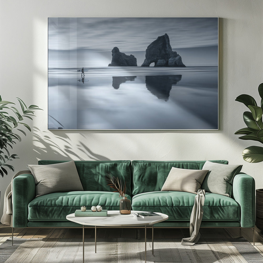 A moody, blue-toned monochromatic photograph of a lone surfer walking on a wet, reflective beach. Two large rock formations stand in the calm ocean in the background, their reflections mirrored in the water. The sky is overcast, creating a serene and atmospheric scene, all enclosed in a silver frame. Decor