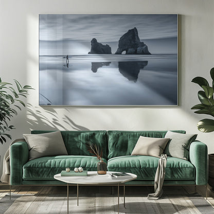 A moody, blue-toned monochromatic photograph of a lone surfer walking on a wet, reflective beach. Two large rock formations stand in the calm ocean in the background, their reflections mirrored in the water. The sky is overcast, creating a serene and atmospheric scene, all enclosed in a silver frame. Decor