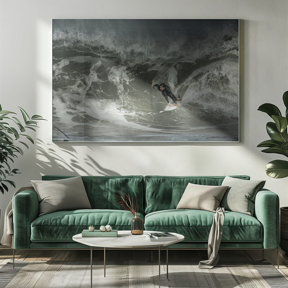 A dramatic overhead photograph of a surfer in a black wetsuit riding a white surfboard on a large, dark, foamy wave. The scene is captured in a moody, almost monochromatic tone and is presented within a silver picture frame. Artwork