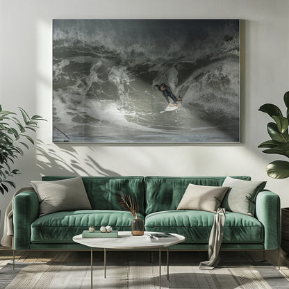 A dramatic overhead photograph of a surfer in a black wetsuit riding a white surfboard on a large, dark, foamy wave. The scene is captured in a moody, almost monochromatic tone and is presented within a silver picture frame. Artwork