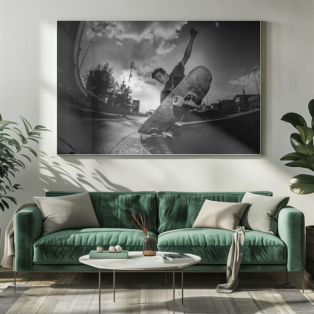 A dynamic, low-angle, black and white photograph of a young man skateboarding. He is balancing on the edge of a ramp with his skateboard angled up towards the viewer. He has one arm raised in the air with a clenched fist. The shot is taken with a fisheye lens, curving the horizon against a dramatic cloudy sky in an urban setting. The image is presented within a silver frame. Decor