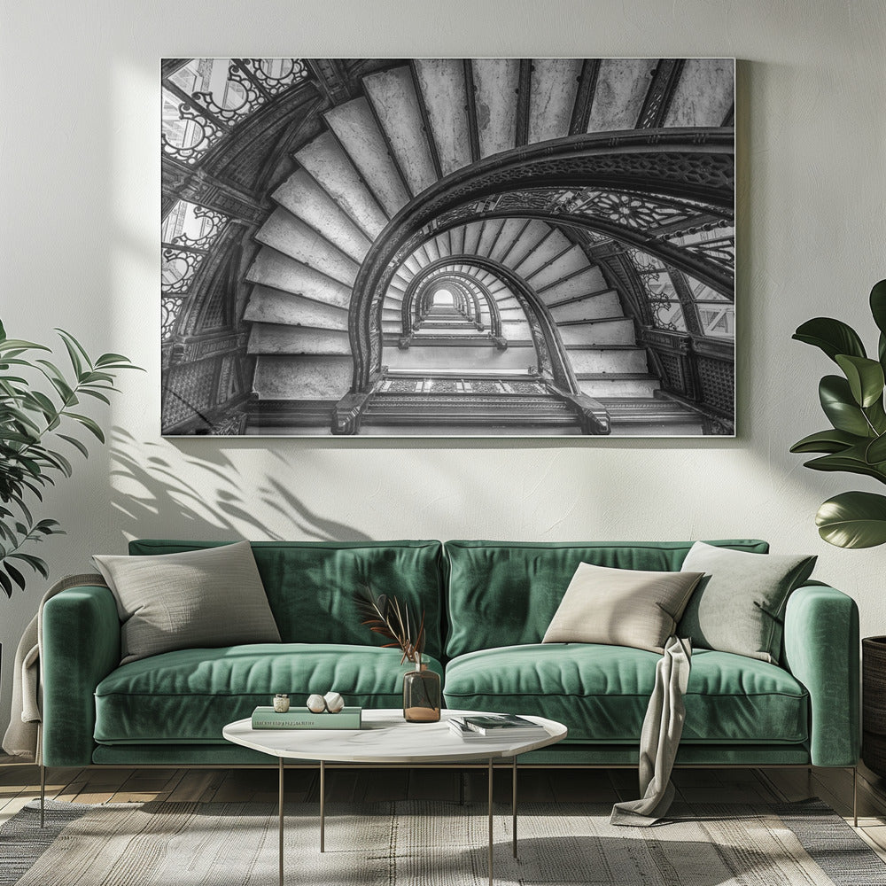 A dramatic black and white photograph looking up from the bottom of an ornate spiral staircase. The dark, intricate ironwork of the railings contrasts with the light stone steps, creating a mesmerizing, repeating pattern that spirals towards a bright light at the top. The image is presented within a silver-colored frame. Decor