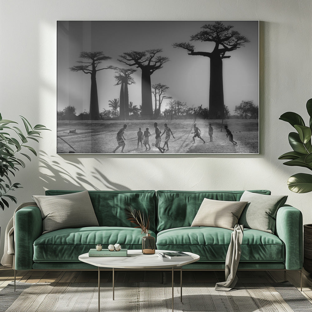 A framed, black and white photograph capturing a vibrant scene of several children playing soccer on a dirt field. In the background, the iconic silhouettes of large Baobab trees stand tall against a bright, hazy sun, casting long shadows of the players across the ground. Artwork