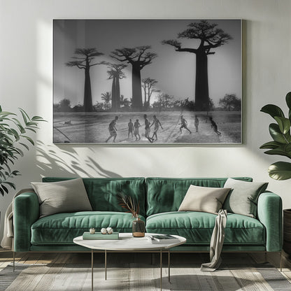 A framed, black and white photograph capturing a vibrant scene of several children playing soccer on a dirt field. In the background, the iconic silhouettes of large Baobab trees stand tall against a bright, hazy sun, casting long shadows of the players across the ground. Artwork