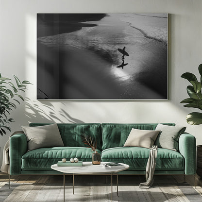A high-contrast, black and white photograph of a silhouetted surfer carrying a surfboard and walking along the wet, reflective sand at the water's edge. Decor