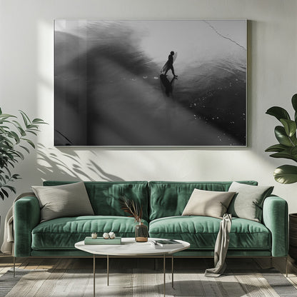 A high-contrast, black and white photograph of a surfer silhouetted against the bright reflection of the sun on the water. The surfer is carrying a surfboard and walking along the wet sand, leaving footprints behind. Their long shadow is cast in front of them on the dark, glistening sand. Print