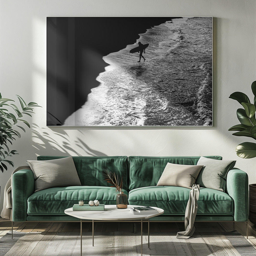 A high-contrast, black and white aerial photograph of a silhouetted surfer carrying a surfboard and walking along the shoreline. The image is divided diagonally, with the dark beach on the left and the white, foamy ocean waves on the right, creating a dramatic and graphic composition. The image is enclosed in a silver frame. Wall Art