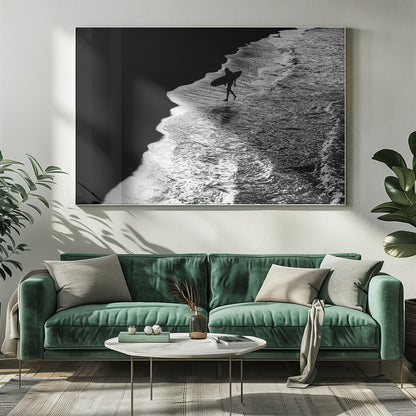 A high-contrast, black and white aerial photograph of a silhouetted surfer carrying a surfboard and walking along the shoreline. The image is divided diagonally, with the dark beach on the left and the white, foamy ocean waves on the right, creating a dramatic and graphic composition. The image is enclosed in a silver frame. Wall Art
