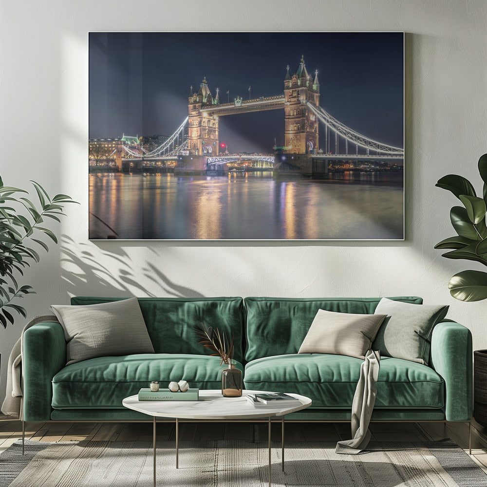 A stunning night photograph of London's iconic Tower Bridge, fully illuminated with golden and white lights that cast a shimmering reflection on the dark waters of the River Thames below, all set against a deep night sky and enclosed in a silver frame. Poster