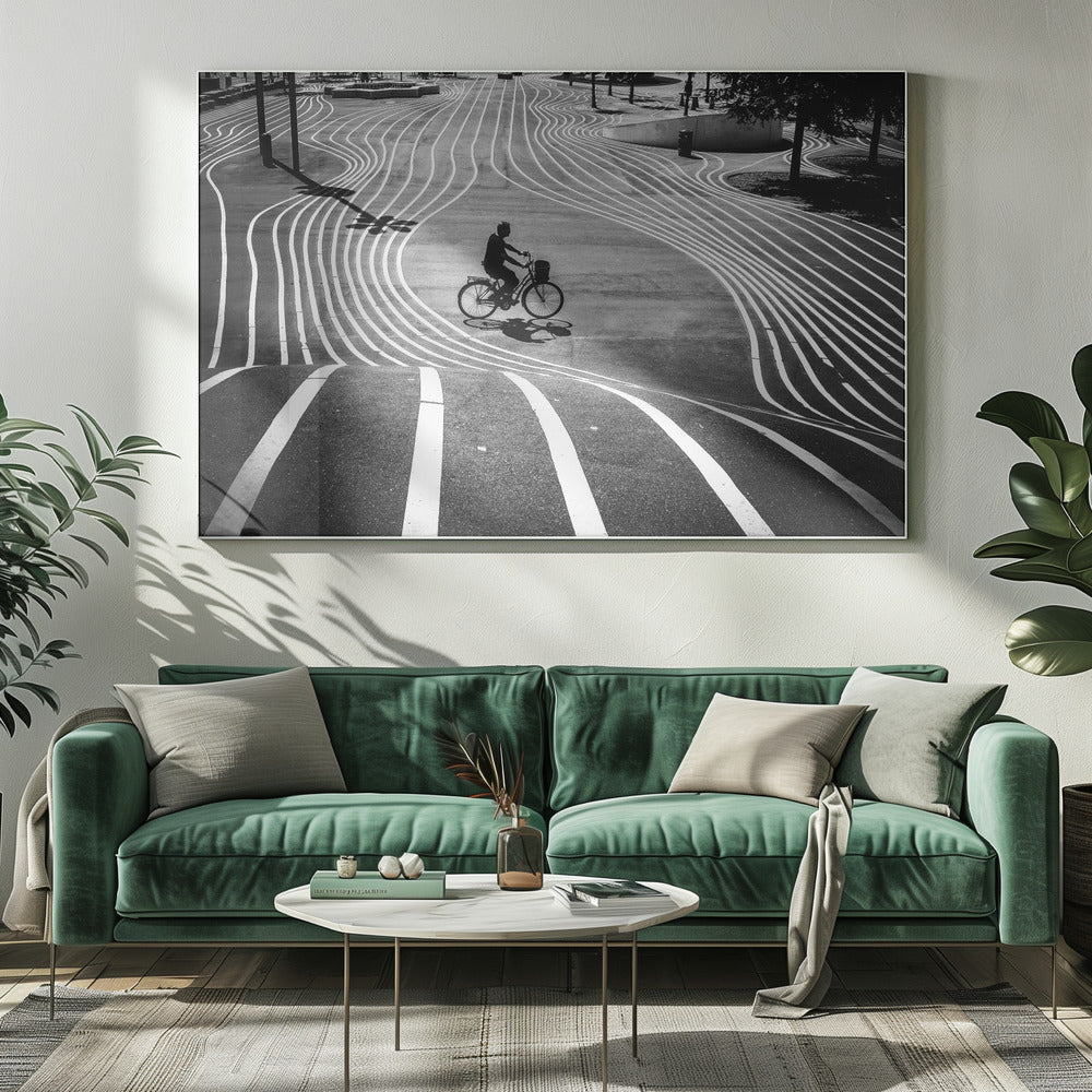 A high-contrast, black and white overhead photograph of a person riding a bicycle across a wide, paved plaza. The ground is covered in a mesmerizing pattern of wavy, parallel white lines, and the cyclist's long shadow is cast by the bright sun. Print