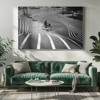 A high-contrast, black and white overhead photograph of a person riding a bicycle across a wide, paved plaza. The ground is covered in a mesmerizing pattern of wavy, parallel white lines, and the cyclist's long shadow is cast by the bright sun. Print