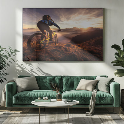 A mountain biker, seen from behind, stops at the top of a grassy hill to admire a spectacular sunset over a vast mountain range. The valleys below are filled with mist, and the sun's golden rays create a stunning starburst effect against the silhouetted peaks. Print