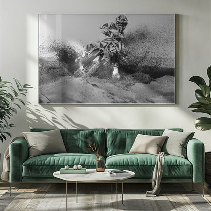A dynamic black and white action photo of a motocross rider in full gear leaning hard into a turn, kicking up a large spray of dirt and sand from the track. The image is presented within a silver frame. Decor